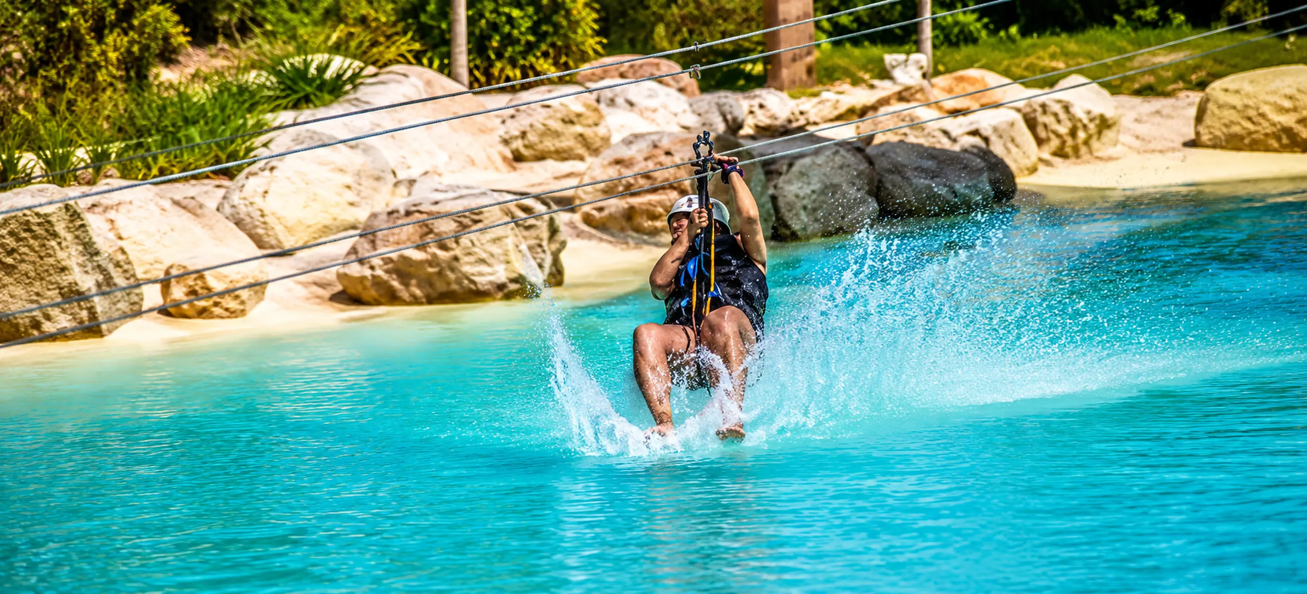 Guest riding the Scape Park Cap Cana water zipline while splashing across a crystal-clear turquoise lagoon during an eco-adventure in Punta Cana, Dominican Republic